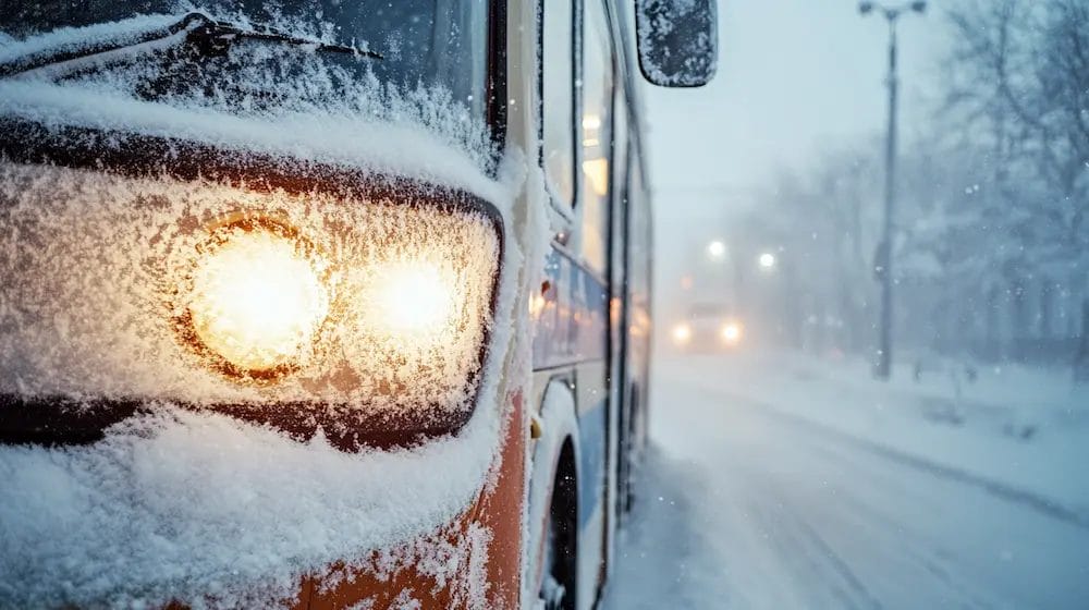 A close-up view of a public transit bus enveloped in a thick layer of snow, showing intricate details like frozen windows and snow-dusted headlights. The contrasting bright colors of the bus peek through the white blanket, creating a striking visual. The environment is quiet and tranquil, embodying the calm of a winter morning.